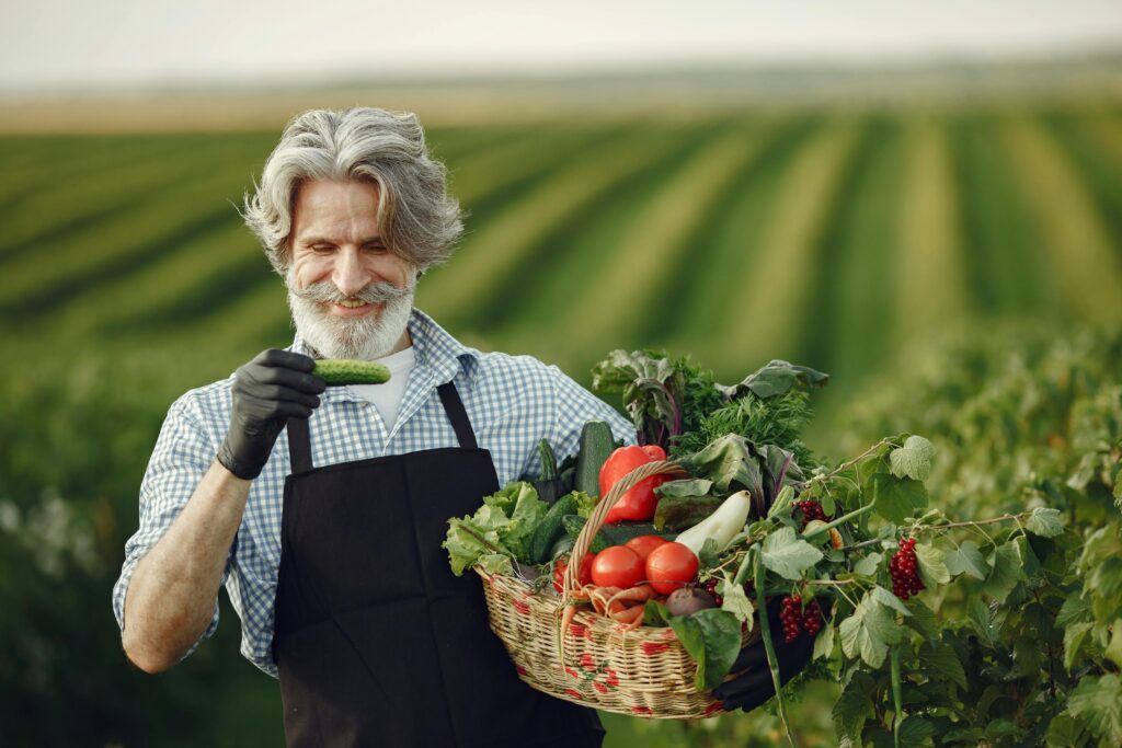 A smiling elderly man holding a basket of fresh vegetables in a lush farm setting during daylight.